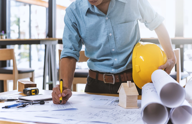 A contractor looking over architectural drawings while holding a hardhat in one hand and a pencil in the other.